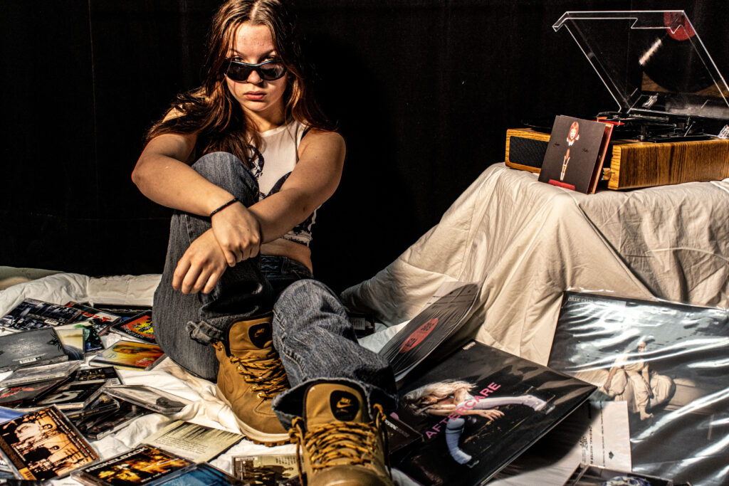 une femme assise dans une pile de cd et vinyles qui fixe la camera